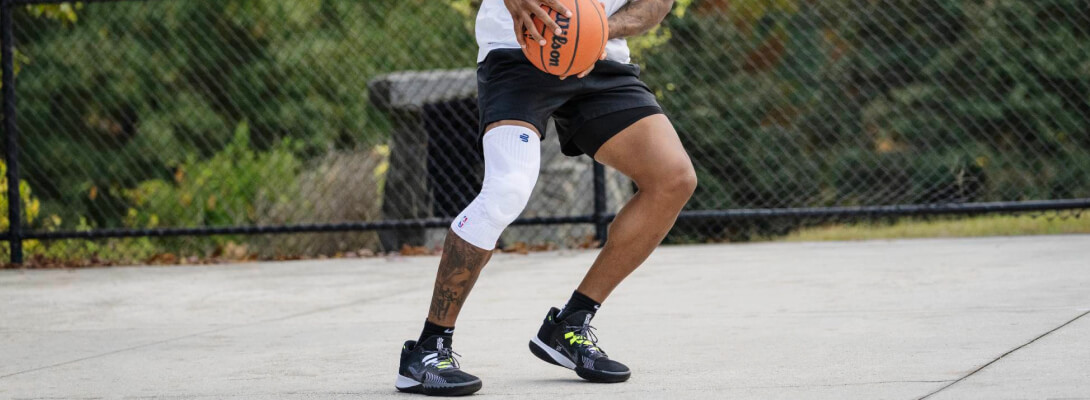 Giocatore con ginocchiera bianca mentre controlla la palla Giocatore di basket con ginocchiera compressiva bianca mentre controlla il pallone su un campo all’aperto. Il tutore si distingue chiaramente e mostra il logo NBA.
