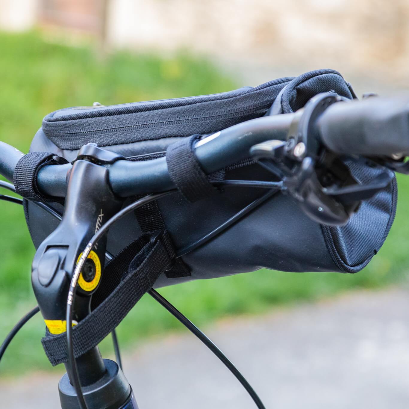 Primo piano della borsa bici nera sul manubrio, con cinghie di fissaggio, leva del freno, cavi e zip superiore ben visibili – la borsa resta compatta sul cockpit e mantiene gli oggetti essenziali a portata di mano durante la pedalata.