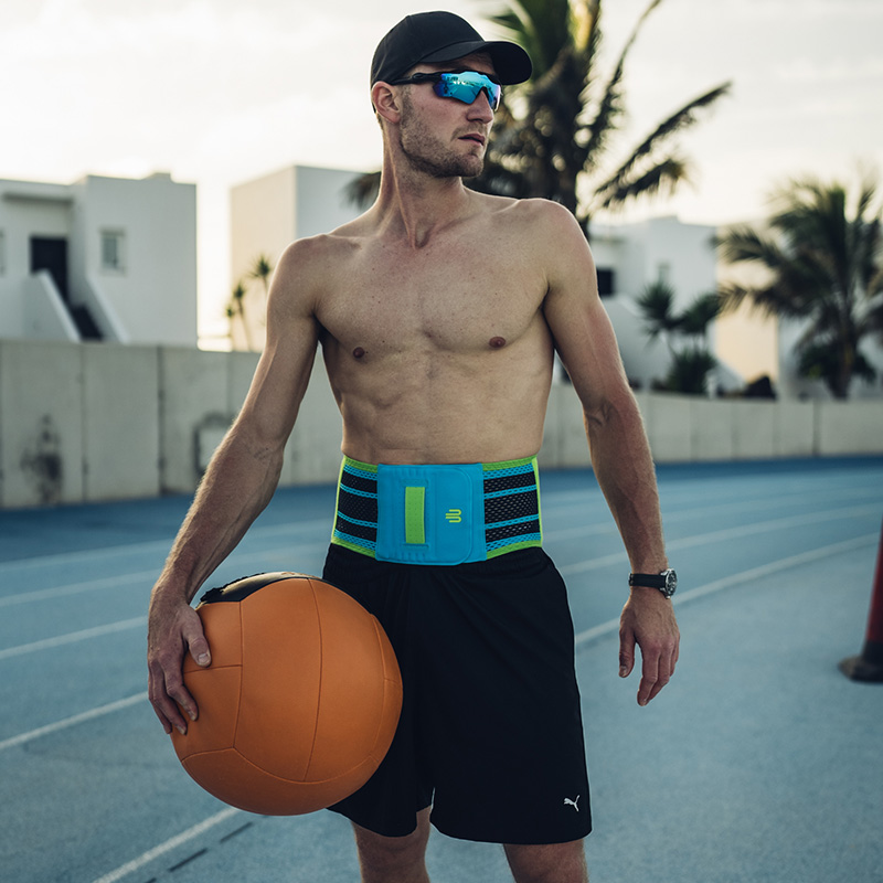 Un uomo sportivo con cappellino e occhiali da sole si trova su una pista da corsa e indossa un supporto lombare blu con accenti verdi. Tiene in mano una grande palla medica.