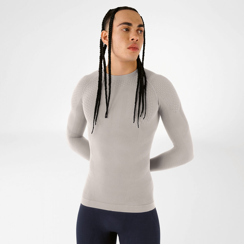 Modello con trecce lunghe indossa una maglia compressiva beige a maniche lunghe davanti a uno sfondo neutro. La superficie strutturata evidenzia il design funzionale senza cuciture.