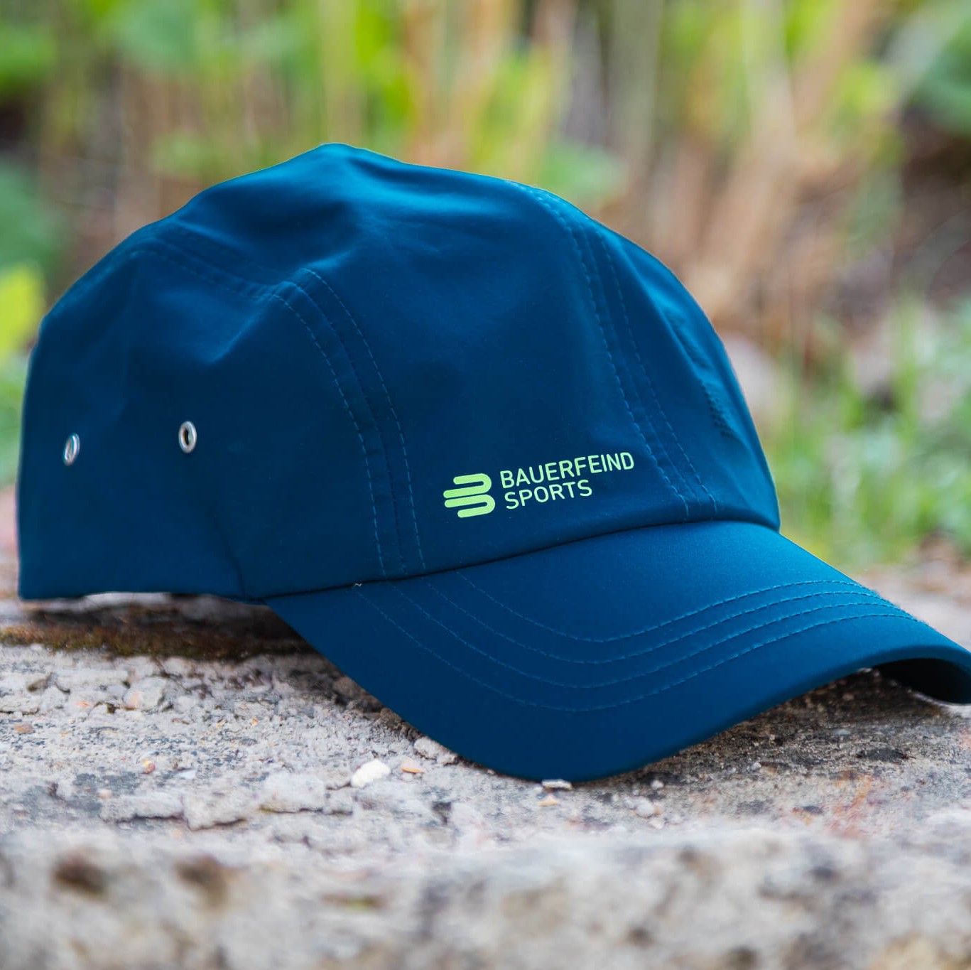 Cappellino blu Bauerfeind Sports con logo verde appoggiato su una pietra all’aperto, circondato da verde sfocato sullo sfondo – un compagno sportivo per le giornate attive outdoor.