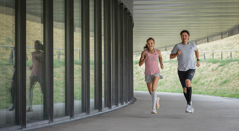 Più forti insieme – chilometro dopo chilometro. Due runner – una donna e un uomo – corrono insieme sotto un passaggio coperto. La donna indossa manicotti a compressione bianchi, l’uomo neri. Entrambi si muovono in sincronia, concentrati e pieni di energia.